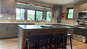 Una cocina moderna con una isla grande, taburetes de bar, electrodomésticos de acero inoxidable y ventanas amplias.