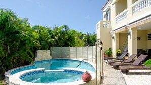Une piscine bordée de pierres, agrémentée d’un vase décoratif, entourée de palmiers et dotée d’un patio couvert avec des chaises longues.