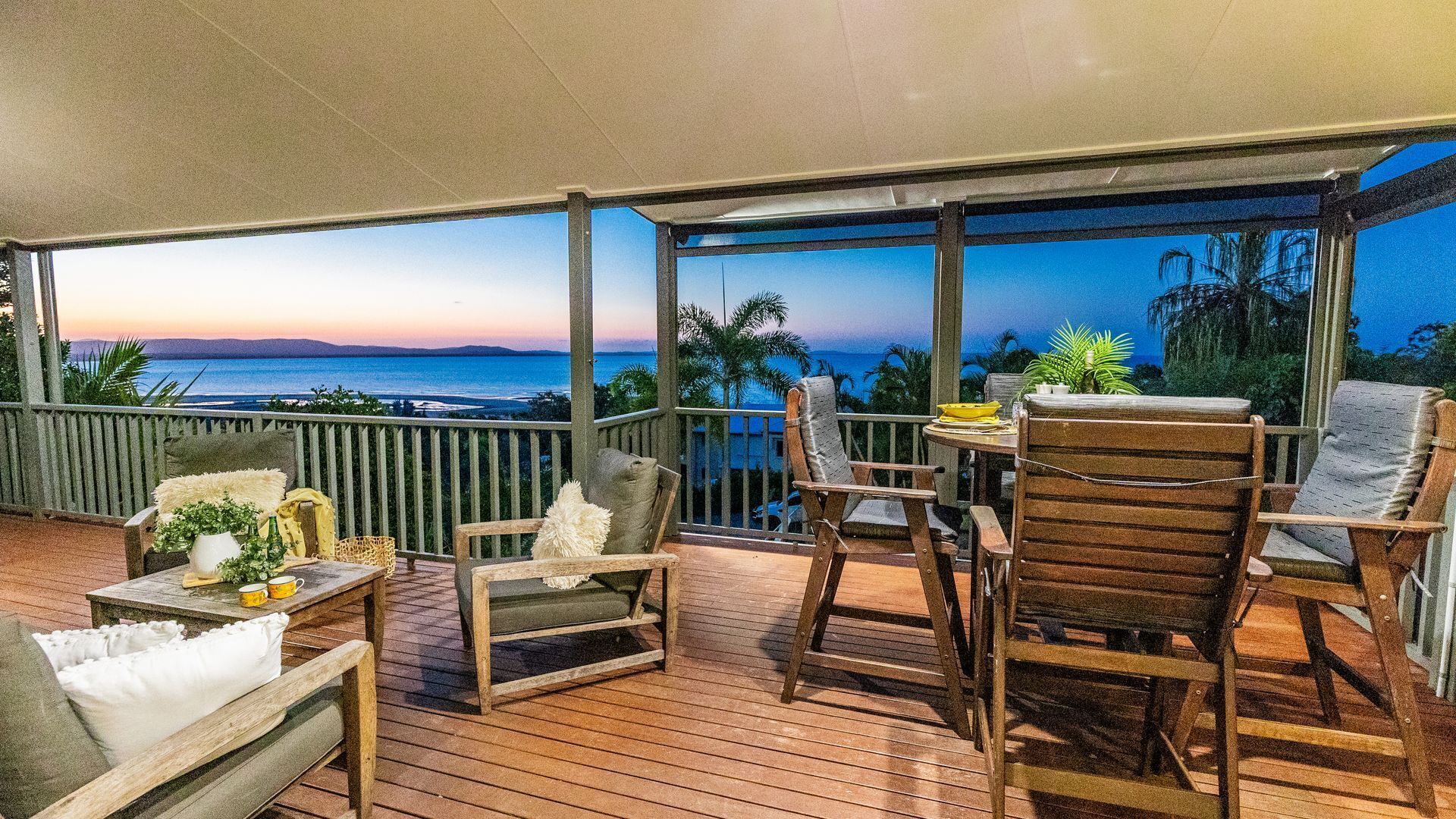 A wooden deck with outdoor furniture, a view of the ocean at sunset, and a covered area with a railing.