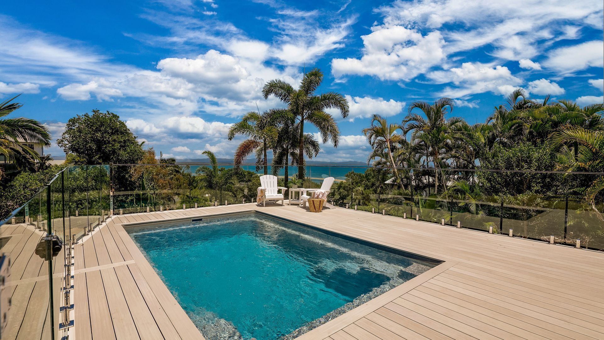 A rectangular pool with a wooden deck, surrounded by palm trees and chairs, overlooking a scenic view.