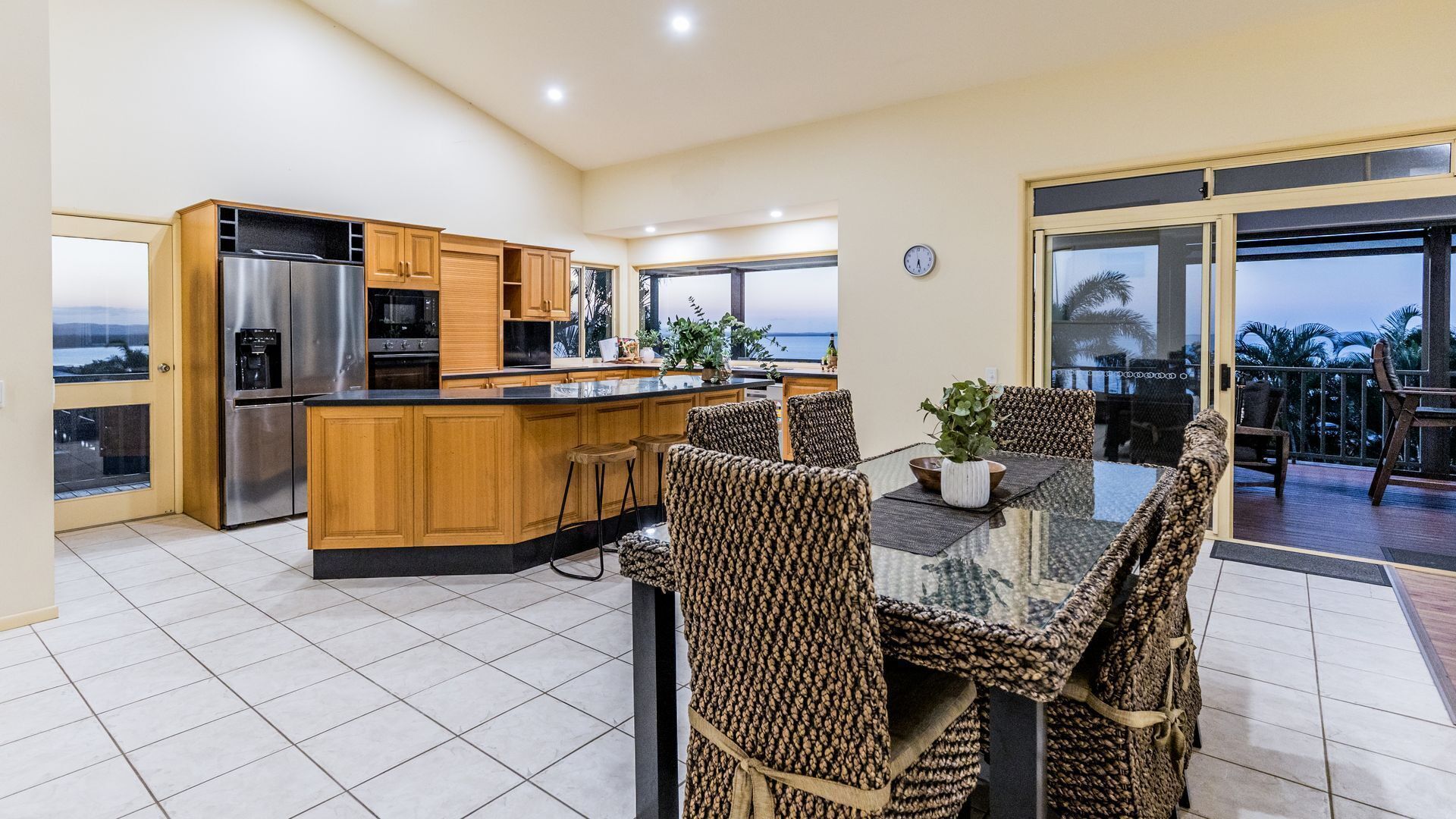 A modern kitchen with a central island, wooden cabinets, and stainless steel appliances. There is a dining area with wicker chairs and a glass table.