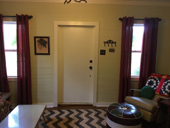 A room with a white door, a framed picture on the wall, a small sign above the door, and a patterned rug.