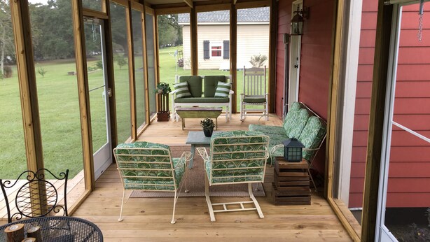 A covered porch with a wooden floor, a green patterned sofa, a rocking chair, and a metal table with a lantern. - Peaceful, Private Cottage On Bayou Teche (New Iberia)