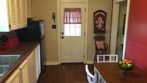 A kitchen with a white door, a window with a pink curtain, a clock, and a fruit bowl on the counter. - Peaceful, Private Cottage On Bayou Teche (New Iberia)