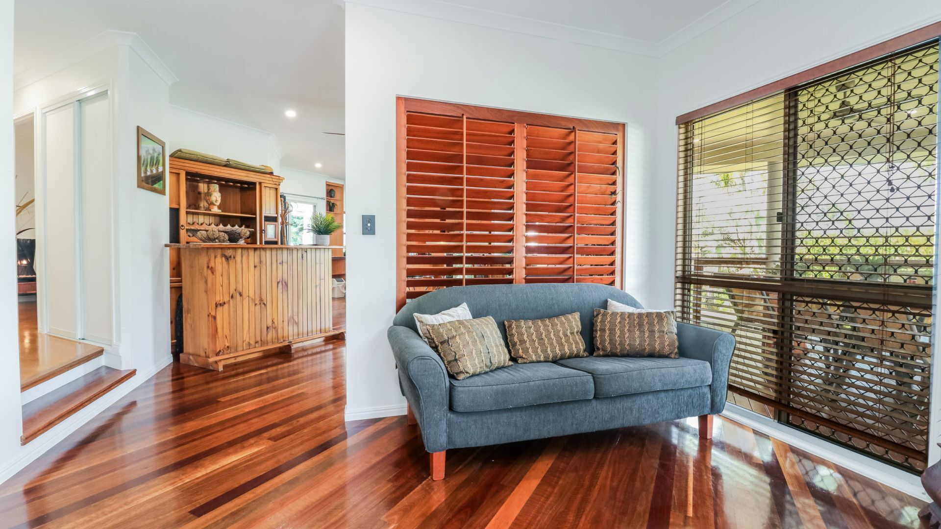 A living room with a grey sofa, wooden cabinet, and a large window with blinds.