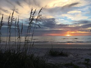 Ein Strand mit hohem Gras im Vordergrund und einem Sonnenuntergang über dem Meer.