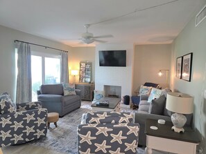 Ein Wohnzimmer mit grauem Sofa, einem Ottomanen mit Meeressternenmuster, einem flachbildfernseher an der Wand, einem Kamin und einem Deckenventilator.