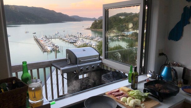 Une cuisine donnant sur un port de plaisance et sur des collines, avec un barbecue, une poêle et divers aliments disposés sur le plan de travail.