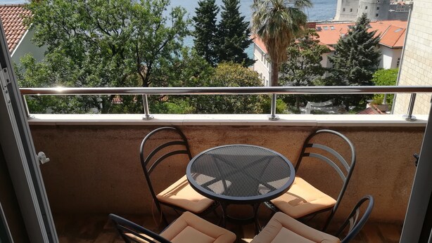 Varanda com mesa e cadeiras, vista para a costa com edifícios e árvores.