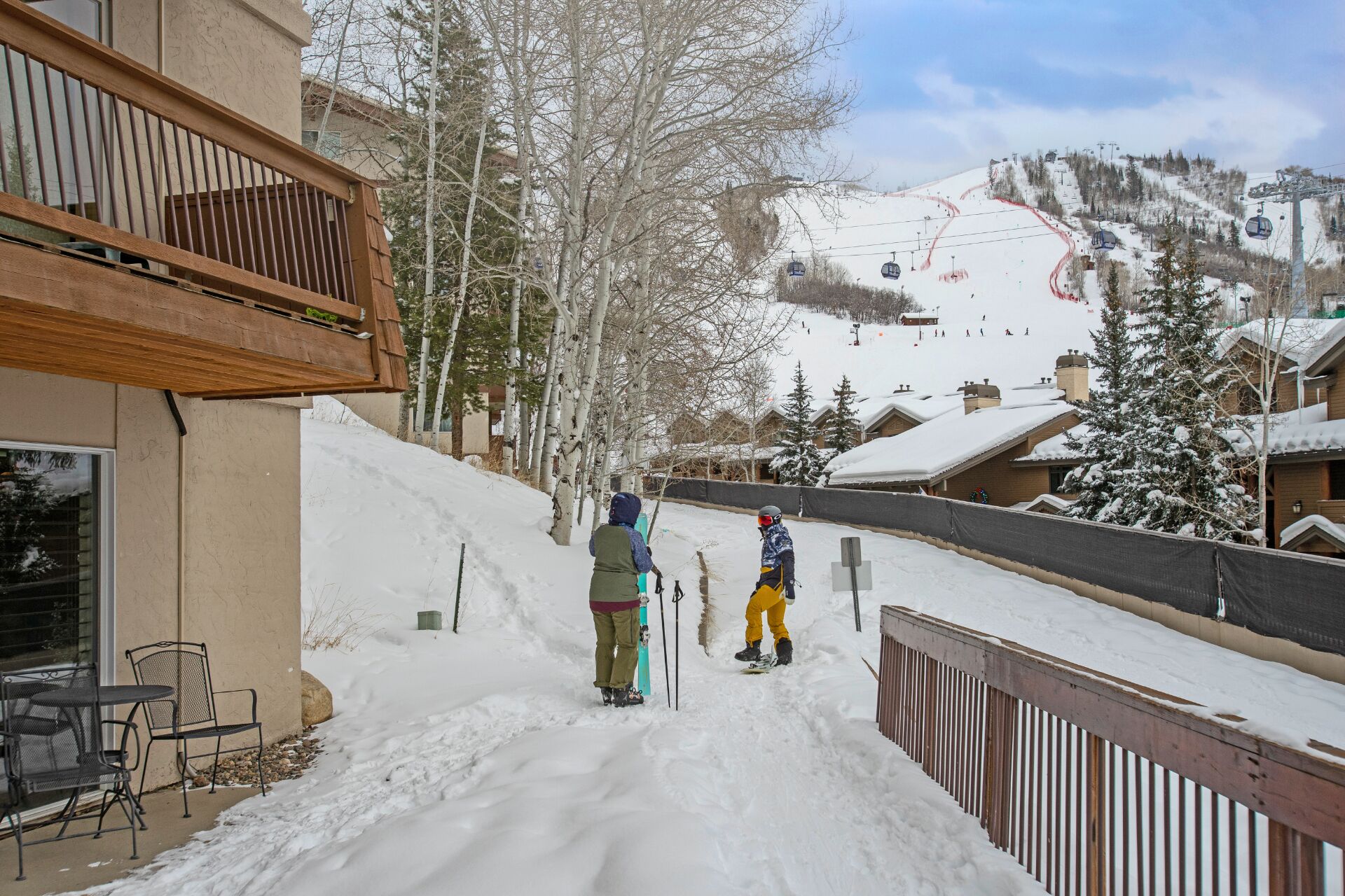 A snow-covered pathway leading to a ski slope with a wooden fence and chairs on the left.