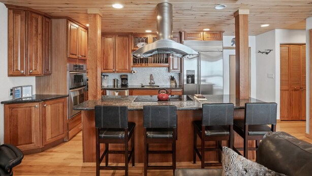 A modern kitchen with wooden cabinets, a stainless steel range hood, a central island with bar stools, and a refrigerator. - Ski In/Out, Walk to Shops, Steam Shower in Unit + Outdoor Hot Tub (Steamboat Springs)
