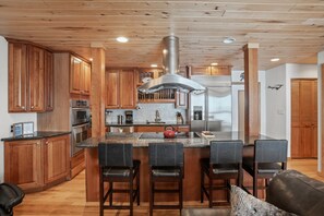 A modern kitchen with wooden cabinets, a stainless steel range hood, a central island with bar stools, and a refrigerator. - Ski In/Out, Walk to Shops, Steam Shower, Hot Tub +EZCancel (Steamboat Springs)