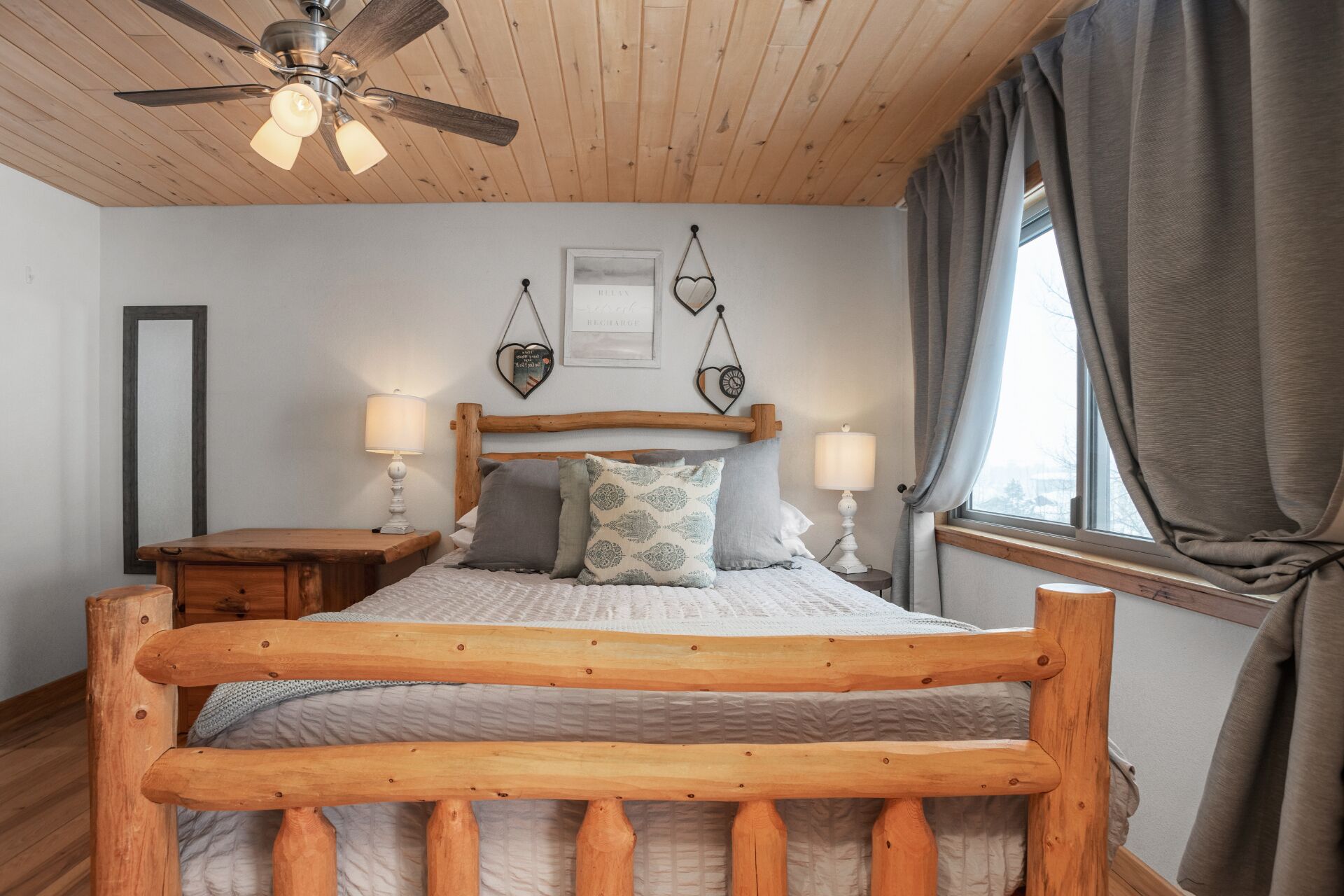 Quarto com estrutura de cama de madeira, duas mesas de cabeceira com luminárias, ventilador de teto e janela com cortinas.