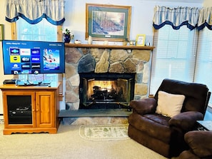 A living room with a stone fireplace, a television displaying a streaming service interface, and a leather armchair.
