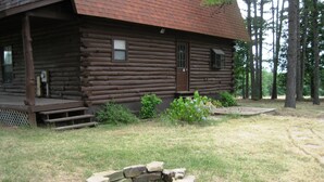 Una baita di legno con un portico anteriore, un braciere in pietra e un'area erbosa circostante con alberi.