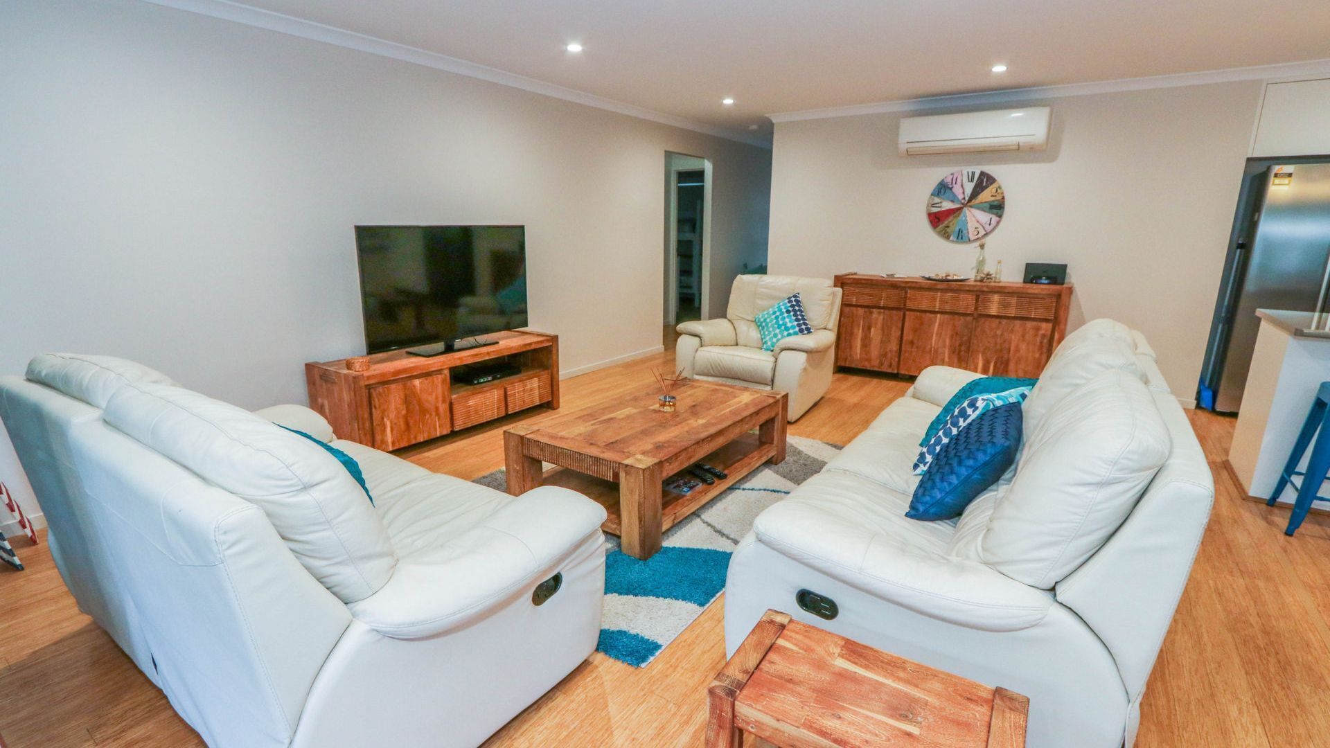 A modern living room with a flat-screen TV, white leather sofas, a wooden coffee table, and a small wooden side table.