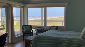 Un dormitorio con una cama grande, una silla, una mesita y vista a la playa a través de amplios ventanales.