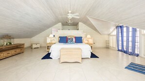 Une chambre spacieuse avec un plafond haut, un grand lit, un ventilateur de plafond et une vue sur l’extérieur par une fenêtre ornée de rideaux bleus.