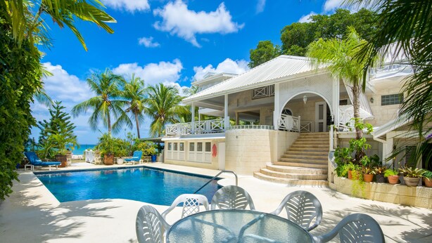 Un espace aménagé au bord de la piscine, avec une table et des chaises en verre, un coin salon couvert et une vue sur l’océan.