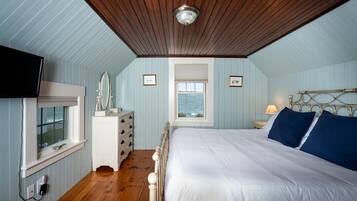 Ein Schlafzimmer mit Holzbalkendecke, einem Bett mit weißer und blauer Bettwäsche, einem Schminktisch, einem Spiegel und einem Fenster mit Meerblick.