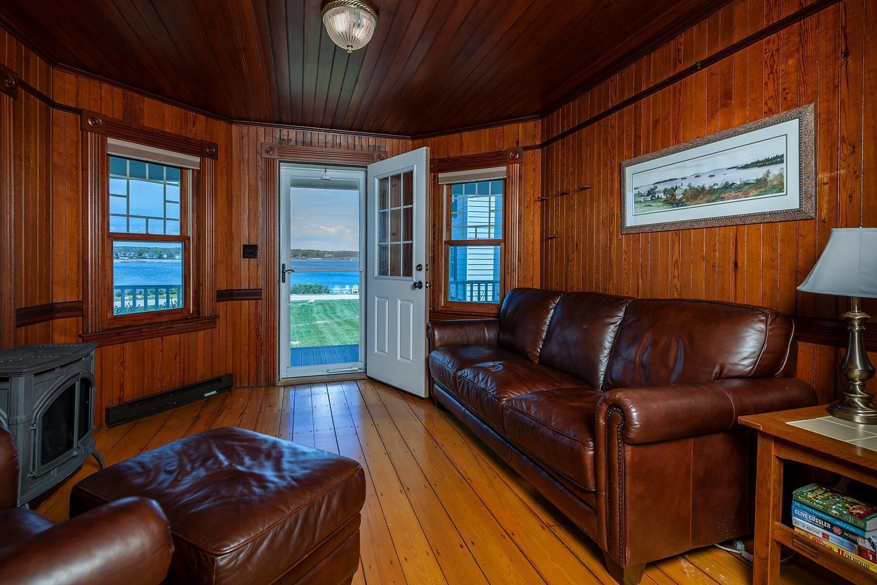 A cozy room with wooden walls, a leather sofa, a fireplace, and a view of the ocean through large windows.