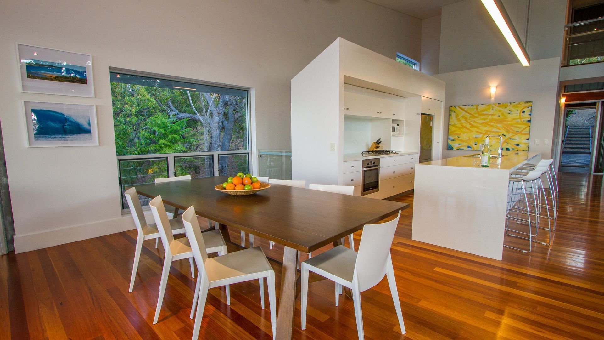 A modern kitchen with white cabinets, a dining area with a wooden table and white chairs, and a view of trees through large windows.