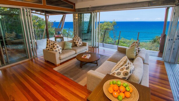 A spacious living area with a wooden floor, a sofa set, a coffee table, and a view of the ocean. - TREE HOUSE - SUNRISE AT 1770 (AGNES WATER)