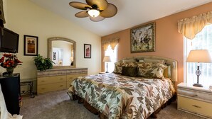 A bedroom with a floral bedspread, a ceiling fan, a dresser, a mirror, and a window with curtains. - Beautiful villa in popular Indian Creek (Kissimmee)