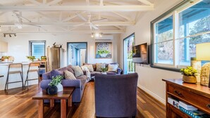 Una sala de estar moderna con sofá, mesa de centro y televisor. Cuenta con una zona de cocina con refrigerador y un taburete de bar.