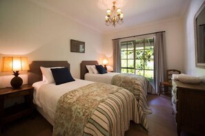 Two beds with headboards and bedside tables, striped bedding, and a decorative throw blanket, set against a backdrop of lush greenery.