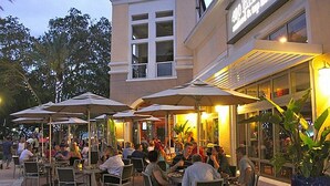 Un espacio al aire libre para comer con mesas y sombrillas, rodeado por un edificio con grandes ventanales y un letrero.