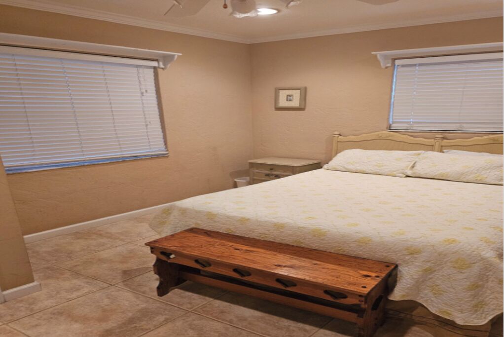 Ein Schlafzimmer mit einem Bett, einem Nachttisch, einer Bank und zwei Fenstern mit Jalousien.