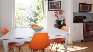 Uma área de refeições com uma mesa branca e cadeiras laranja, uma janela com vista para árvores e uma cozinha com armários brancos e micro-ondas.