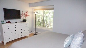 Um quarto com uma cama grande, uma cômoda com várias gavetas, uma TV de tela plana, um vaso de planta e uma janela com vista para árvores.