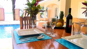 A dining table set with plates, cutlery, and glasses, near a poolside area with a tropical plant and a bottle of wine.