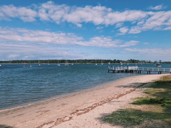 A beach with a wooden pier extending into calm waters, surrounded by boats and a forested shoreline.