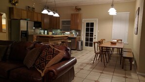Uma sala de estar ampla com um sofá de couro, uma área de jantar com cadeiras de madeira e uma cozinha com armários de madeira e uma geladeira.