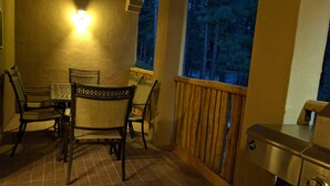 Varanda com mesa, cadeiras, churrasqueira e vista para árvores à noite.