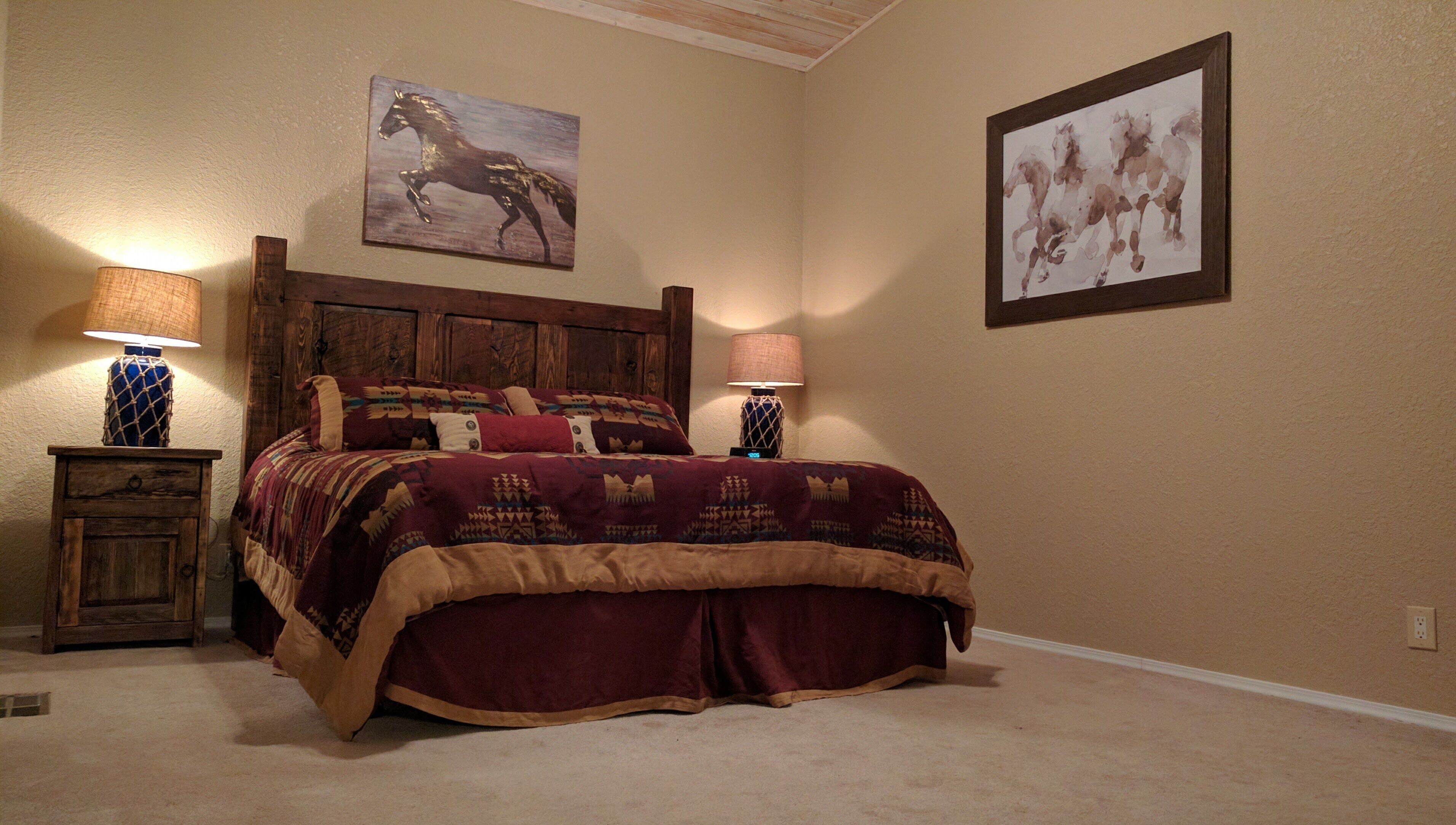 A bedroom with a wooden headboard, a large bed with a patterned bedspread, two bedside tables with lamps, and two wall-mounted paintings.