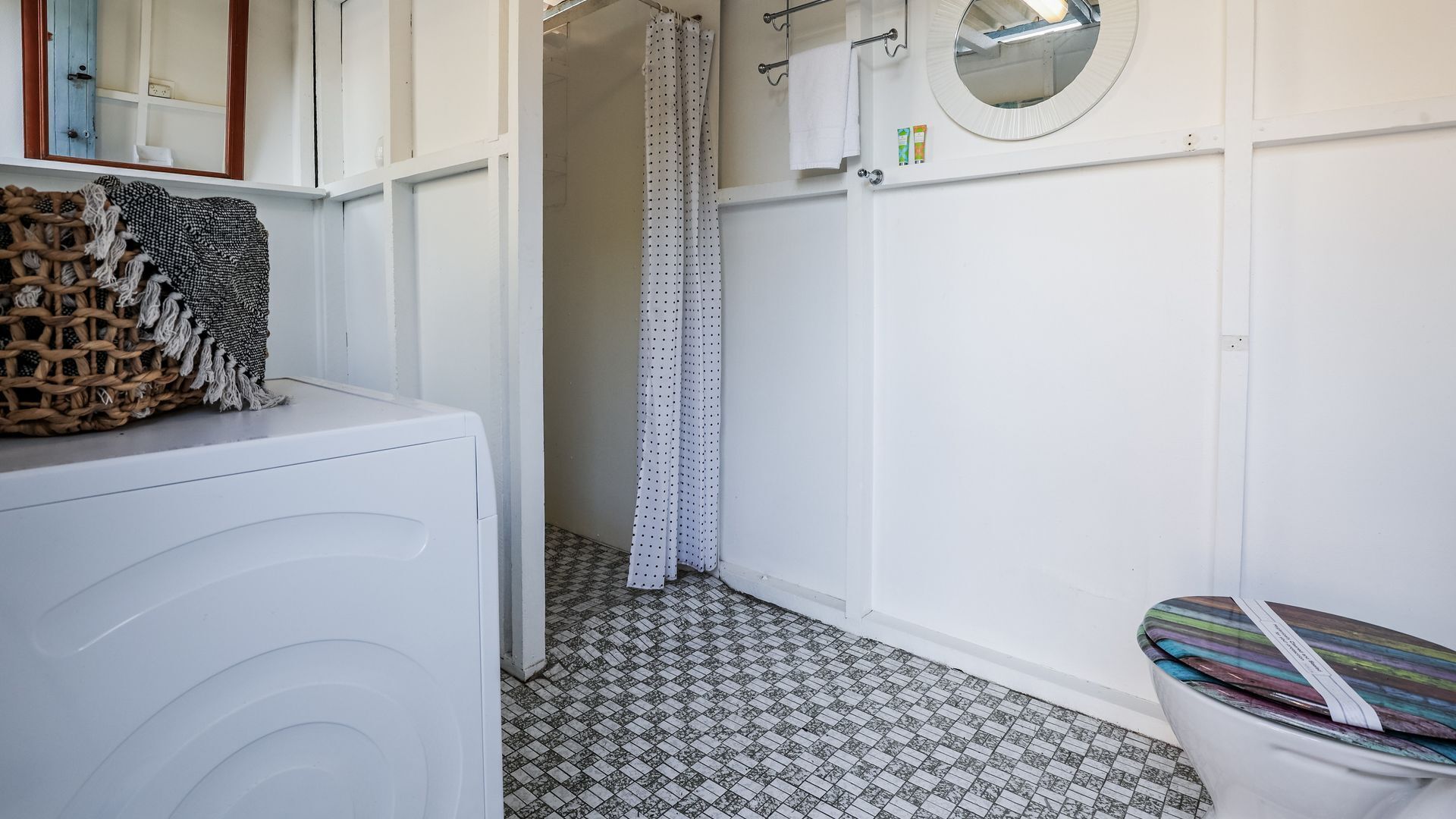 Een badkamer met een witte wasmachine, een douche met een gordijn met stippen, een ronde spiegel en een betegelde vloer.