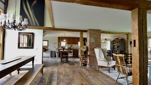 A rustic interior with exposed wooden beams, a dining area with a wooden table and benches, a brick fireplace, and a kitchen in the background. - Long View Farm on Broad Bay (Waldoboro)