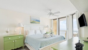 Un dormitorio con cama, mesitas de noche, escritorio, ventilador de techo y vista al océano.
