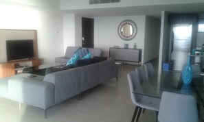 Uma sala de estar moderna com sofá cinzento, televisão de tela plana, mesa de jantar com cadeiras e área de cozinha com geladeira.