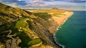 Vista aérea de un paisaje costero con un campo de golf, una playa y montañas al fondo.