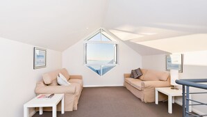 Een knus vertrek met twee beige banken, een witte salontafel en een klein bureau met lamp. Een groot raam biedt uitzicht op de lucht en een verafgelegen landschap.