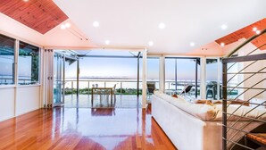 Een ruime kamer met houten vloer, een witte bank en uitzicht op zee door grote ramen.