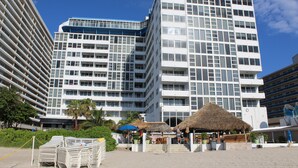 Ein modernes Hotel mit mehreren Etagen, Balkonen und einer Strandlage mit reetgedeckten Sonnenschirmen.