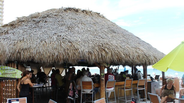 Ein Außenbereich mit Essmöbeln unter Reetdächern, eine Bar und Blick aufs Meer.