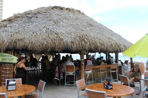 An outdoor dining area with thatched-roof tables and chairs, a bar, and a view of the ocean.