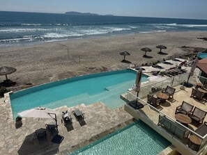 A pool area with lounge chairs and umbrellas, a beach in the background, and a clear view of the ocean. - Stunning, Relaxing Beachfront With Endless Activities & Enjoyment (Rosarito)